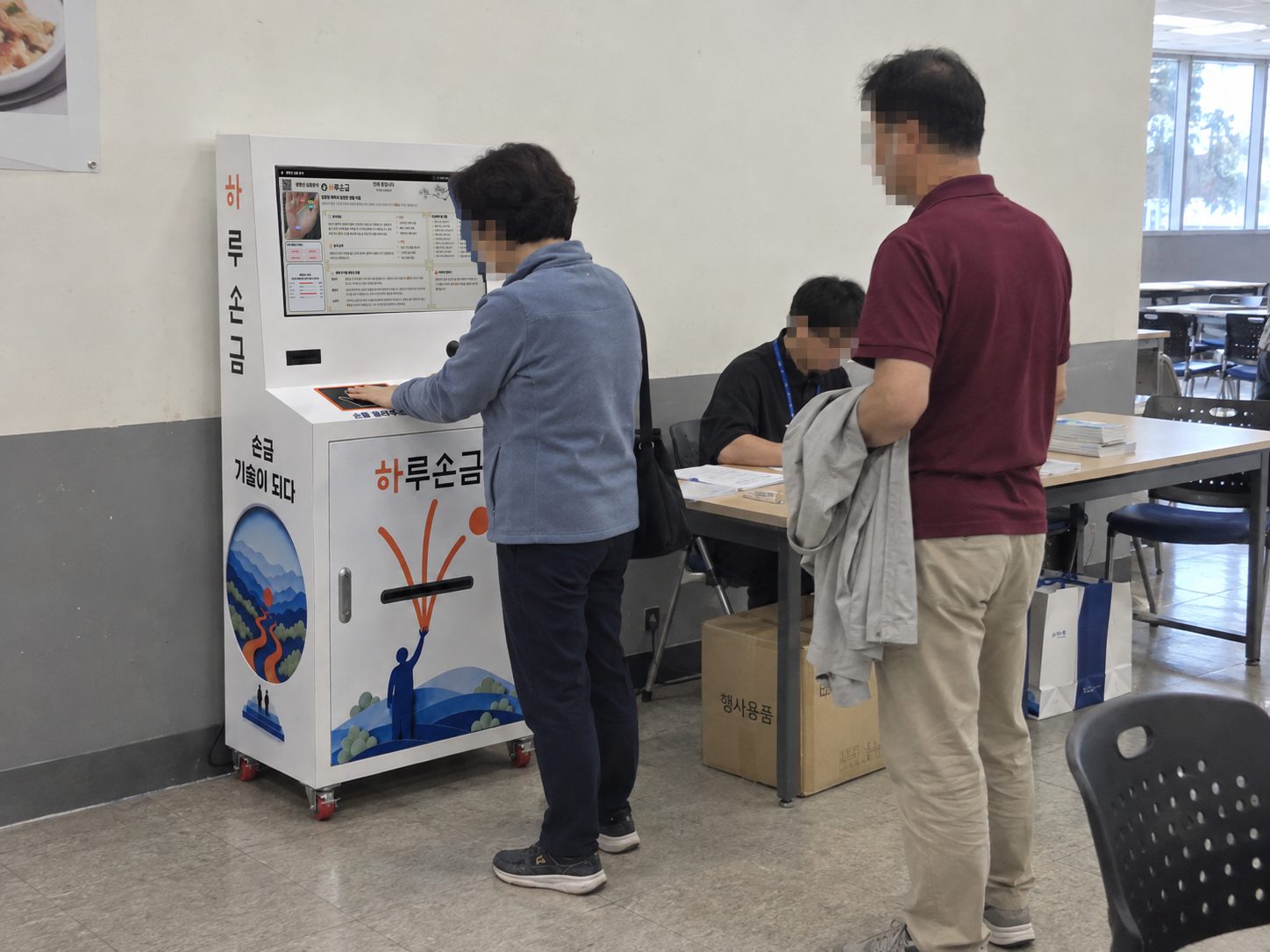 행사 접수대 옆에서 참가자가 하루손금 자판기를 이용하고 접수 담당자가 옆에 앉아 있는 장면